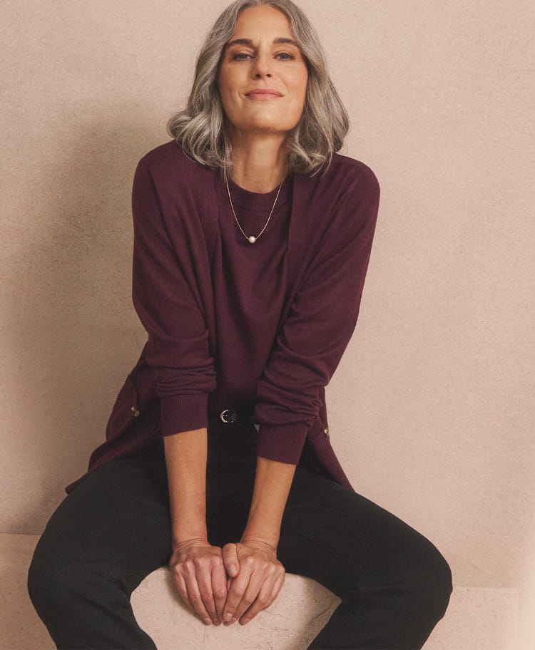 A person is seated against a light beige wall, wearing a long-sleeved maroon top paired with black trousers.