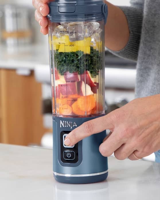 A person pressing the start button on a Ninja blender filled with layered fruits, vegetables, and ice.