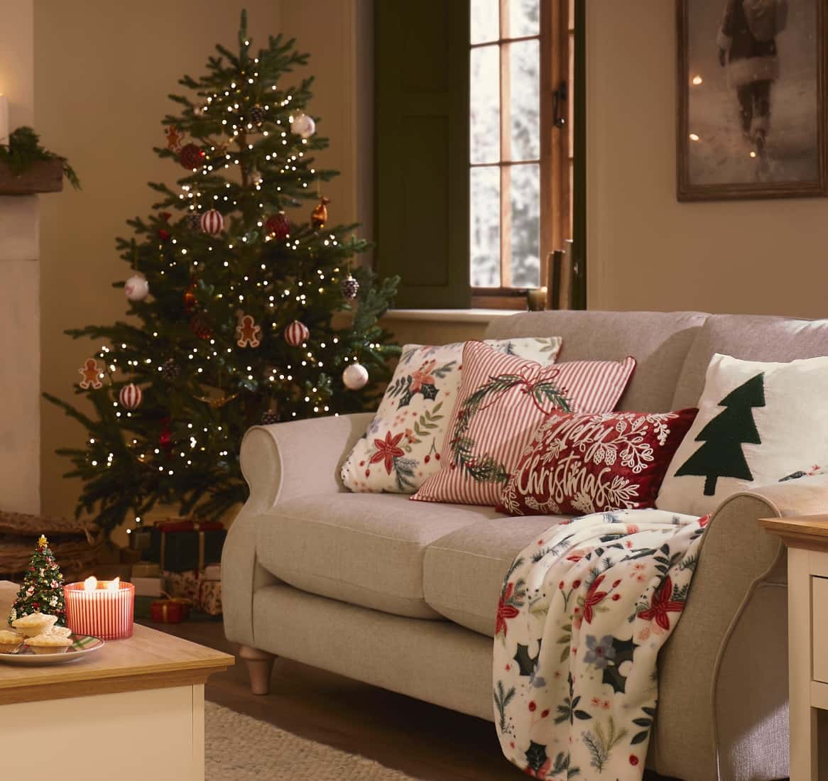 A living room with a decorated Christmas tree, a light grey sofa with festive cushions and throw, and a coffee table with candles and treats.